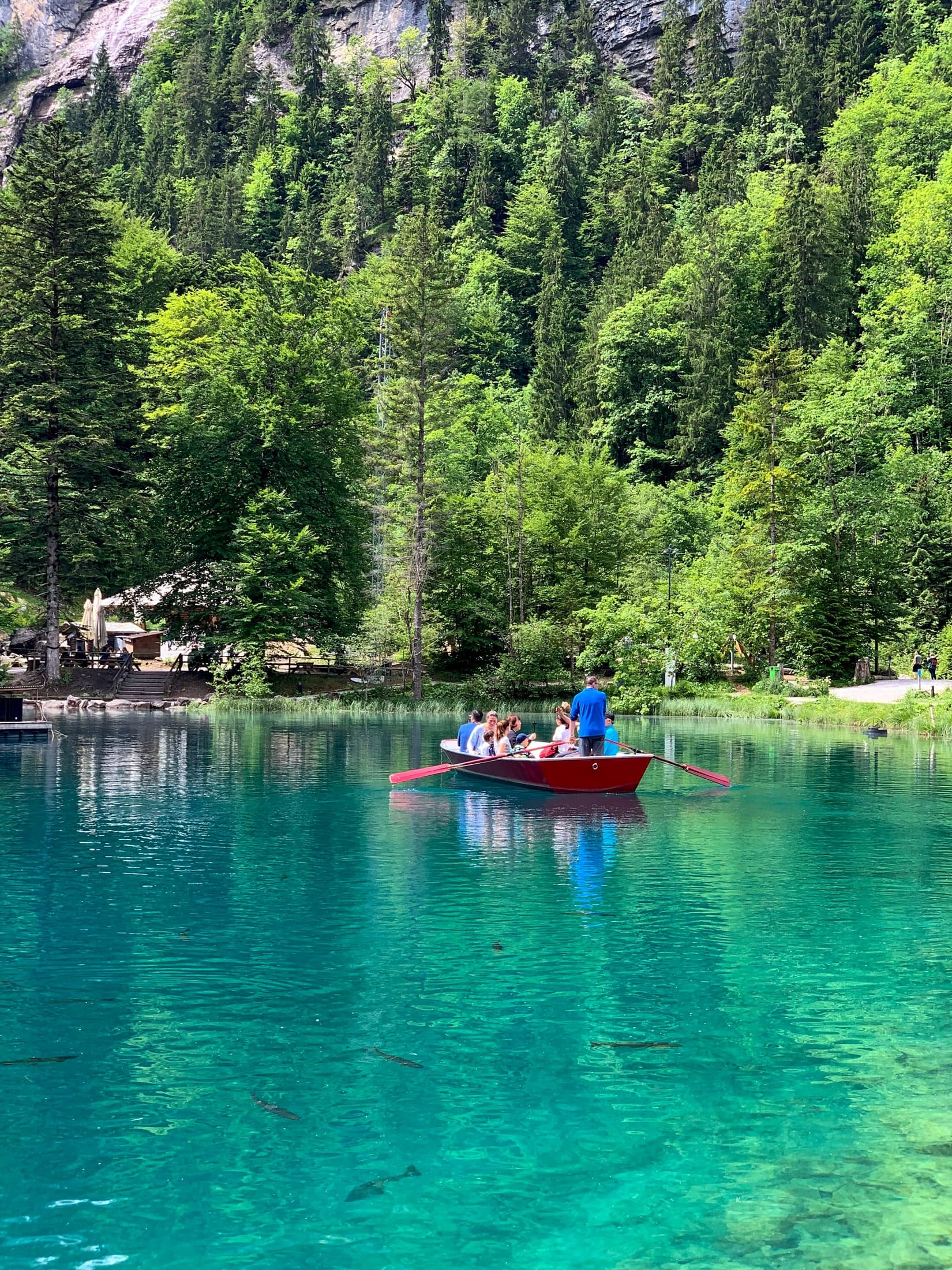 Barco a remo vermelho com pessoas num lago azul-turquesa transparente, cercado de pinheiros