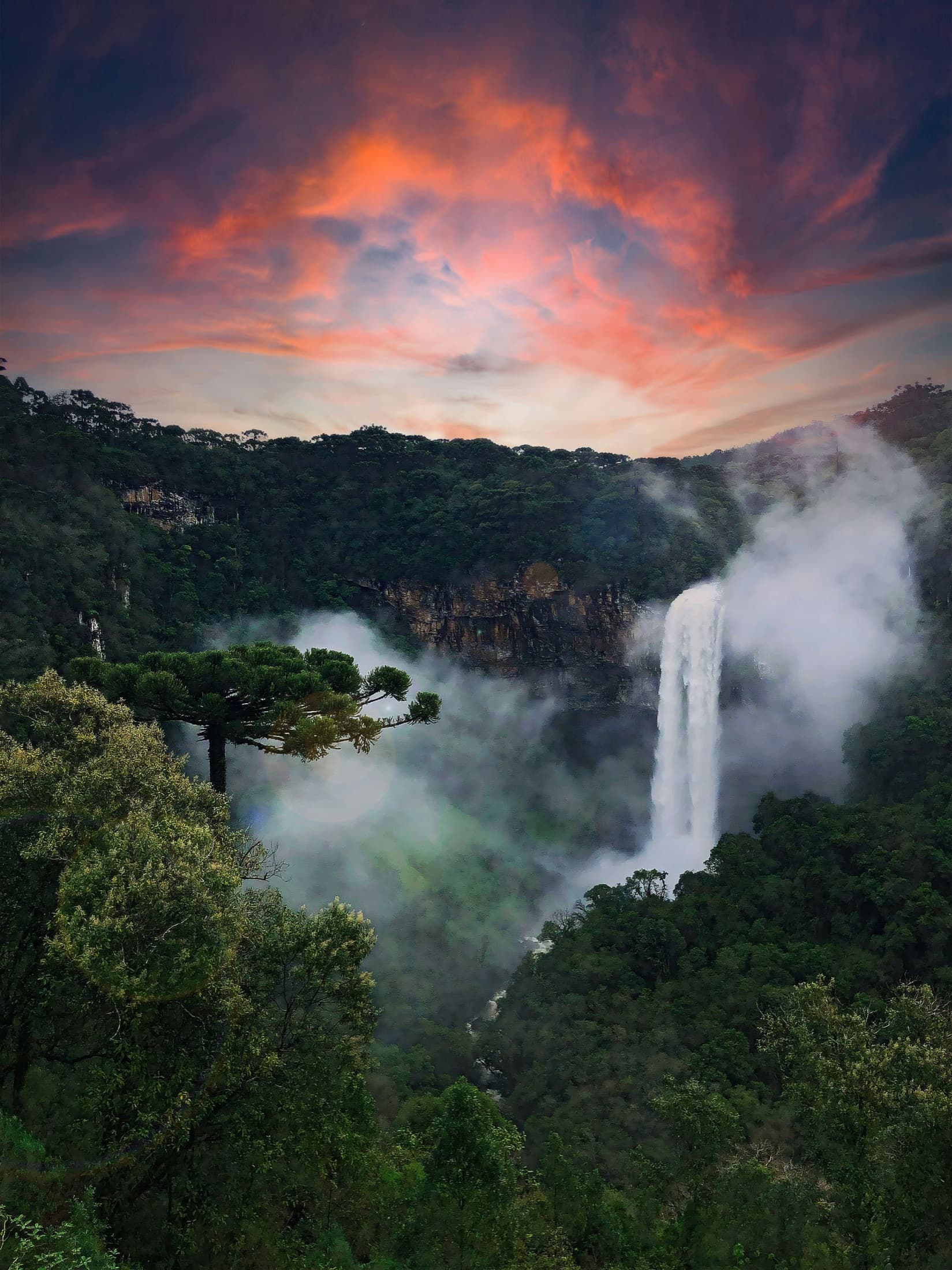 Cachoeira alta despencando por um cânion de mata ao entardecer, sob um céu alaranjado
