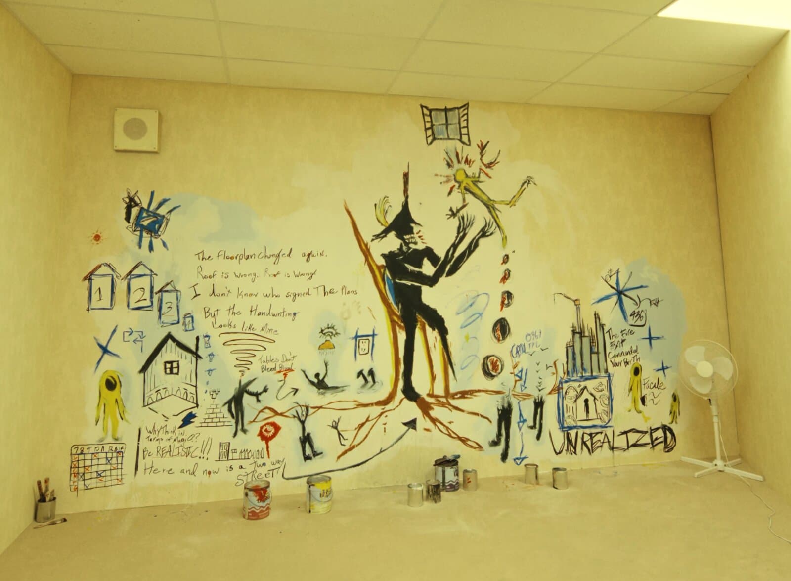 Mural pintado numa parede amarela, no estilo fragmentado de Basquiat: uma figura negra e alongada de chapéu de pirata no centro, textos manuscritos em inglês, casas numeradas, uma janela pintada no alto, a palavra UNREALIZED em letras grandes e pequenas figuras amarelas nas bordas. No chão, latas de tinta abertas; à direita, um ventilador de pé.