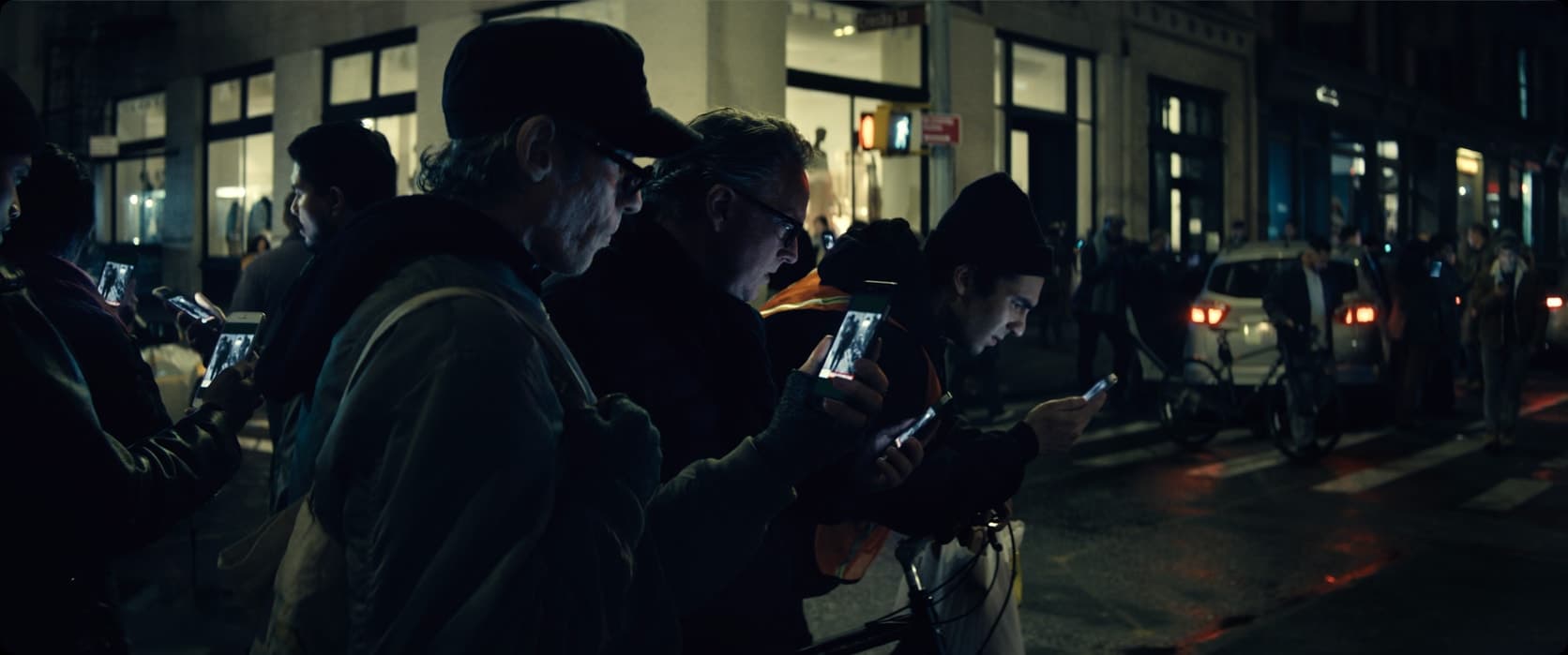 Numa calçada à noite, várias pessoas enfileiradas seguram celulares diante do rosto, todas com a mesma imagem na tela. Vitrines acesas e carros ao fundo.