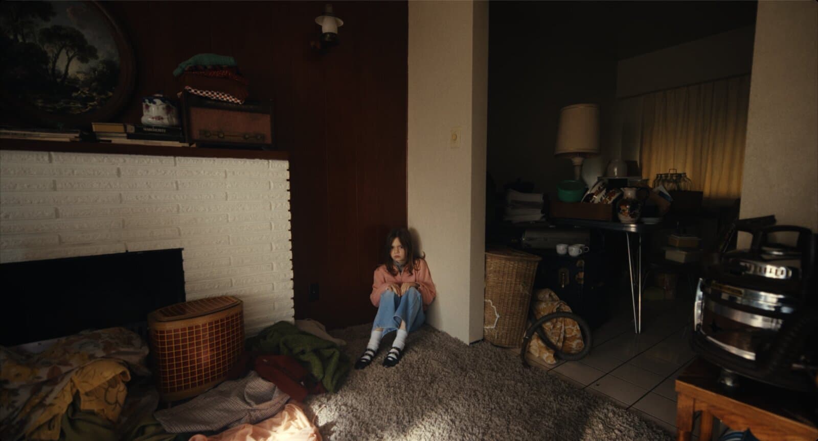 Uma menina sentada no chão, encolhida com os braços em volta dos joelhos, num canto de sala apertado entre uma lareira e uma parede. Ao redor, móveis, cestos, roupas e objetos empilhados por todo o cômodo.