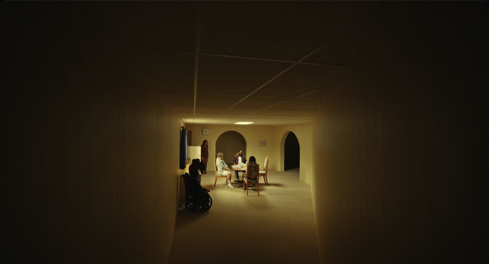 Vista de dentro de um corredor escuro e comprido. Ao fundo, um trecho iluminado com uma mesa de jantar posta, quatro figuras sentadas ou de pé em volta dela, uma geladeira e arcos nas paredes, como se um cômodo doméstico tivesse sido encaixado no meio do corredor.