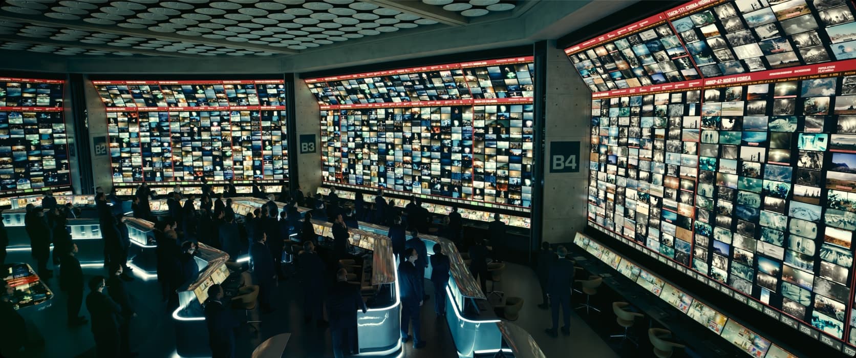 Vista de cima de uma sala de controle ampla. Dezenas de pessoas de terno, em pé entre consoles iluminados, diante de paredes cobertas do chão ao teto por centenas de telas pequenas exibindo transmissões distintas.