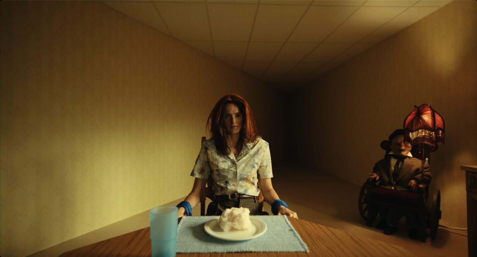 Uma mulher amarrada a uma cadeira com cordas azuis, sentada a uma mesa, usando uma peruca ruiva mal ajustada sobre o próprio cabelo. Diante dela, um prato com uma massa branca e esponjosa. Ao fundo, à direita, uma figura numa cadeira de rodas com um abajur bordô aceso acoplado.