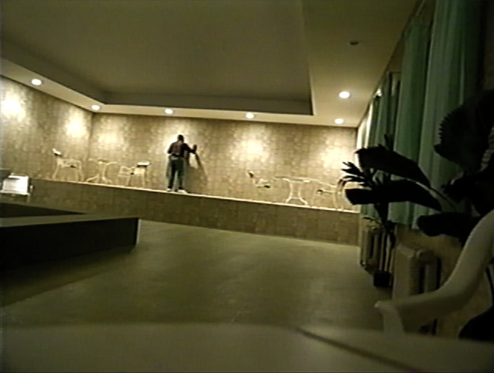 Imagem granulada, com aparência de fita VHS, de um salão amplo e vazio: um homem sozinho está de pé sobre um patamar elevado, de costas para a câmera, com as mãos apoiadas na parede à sua frente. Ao redor dele, cadeiras e mesas brancas de jardim; à direita, uma planta e cortinas.