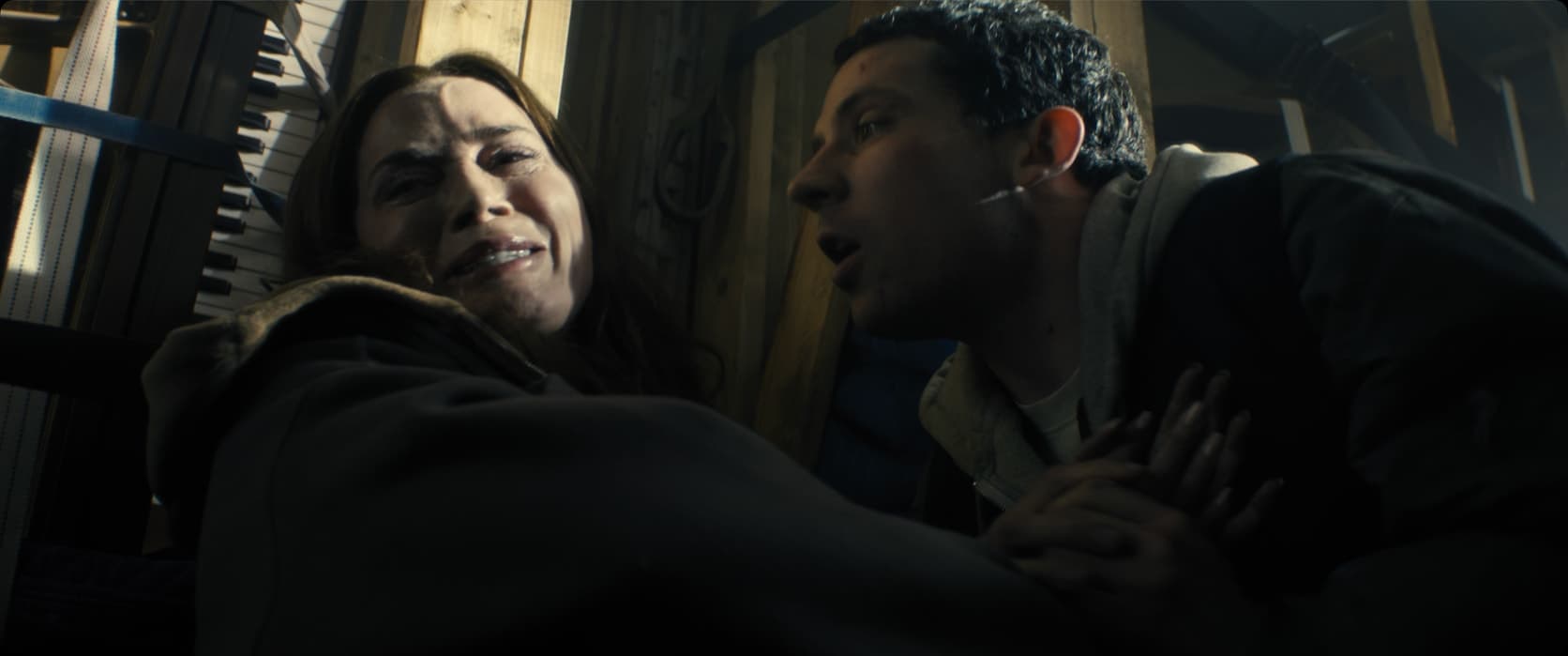 Close de uma mulher caída no chão ao lado de um piano encaixotado, com o rosto contraído e virado para cima, longe do homem ao lado dela. Ele, de moletom com capuz, está inclinado bem perto, falando, com as mãos sobre as dela.