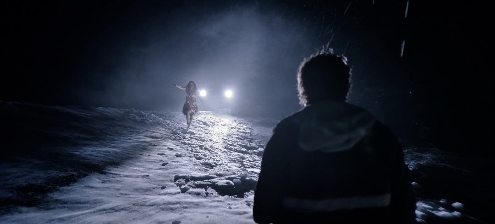 Homem visto de costas, à noite, numa estrada coberta de neve, diante de uma figura de braços estendidos recortada contra dois faróis acesos na névoa