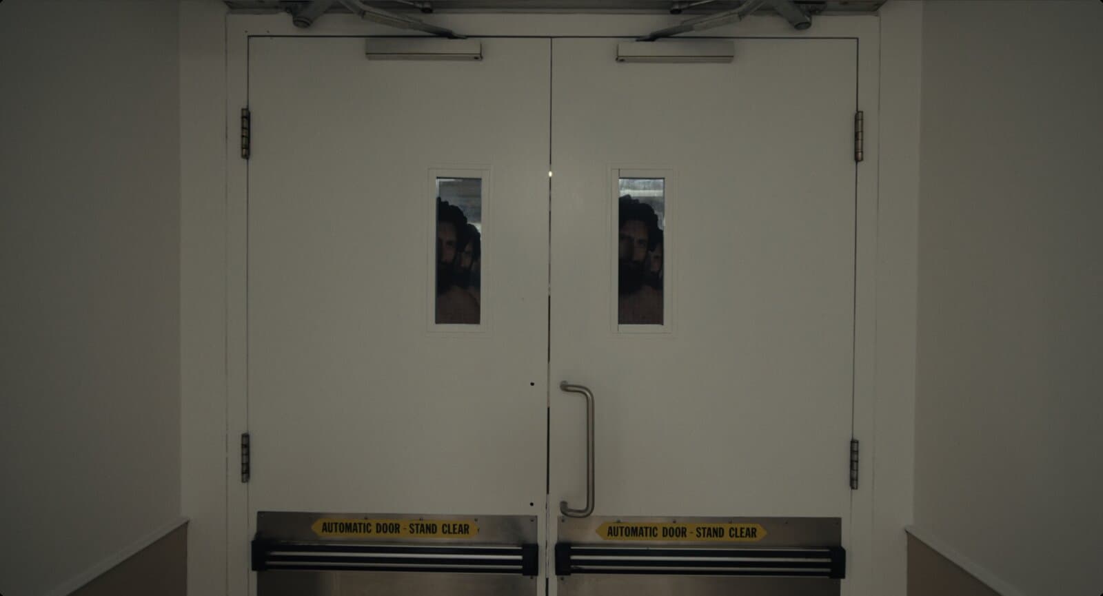 Um par de portas brancas de acesso restrito, com dois pequenos vãos de vidro. Através deles, veem-se recortes de papelão do homem das cavernas enfileirados, um atrás do outro, praticamente idênticos.