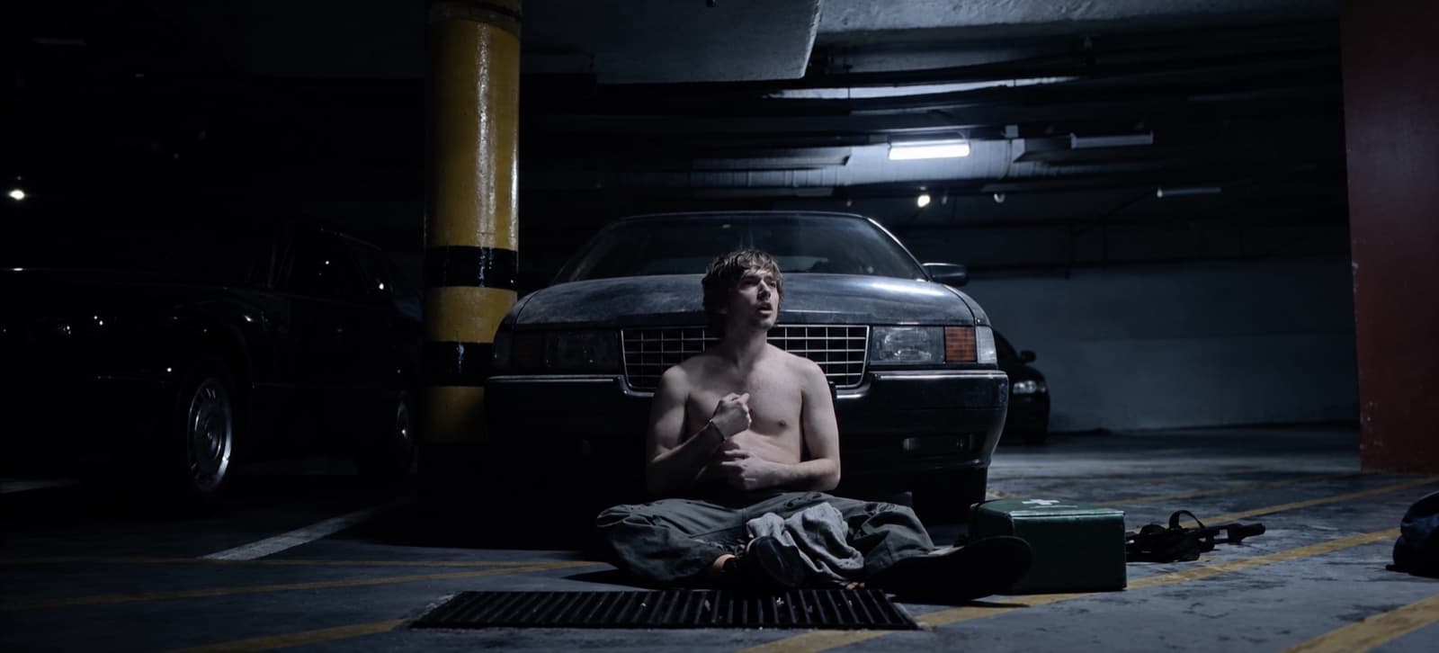 Homem jovem sem camisa sentado no chão de um estacionamento subterrâneo, encostado na frente de um carro, olhando para cima com a mão junto ao peito