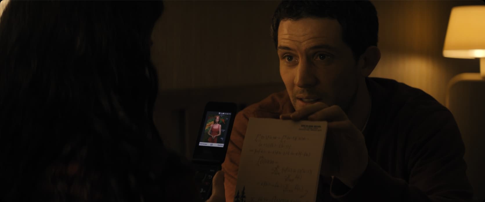 Um homem de cabelo escuro segura junto ao rosto uma folha coberta de equações manuscritas. Diante dele, uma mão estende um celular de tampa cuja tela mostra uma mulher de vestido vermelho falando em pé. Interior escuro, com um abajur aceso ao fundo.