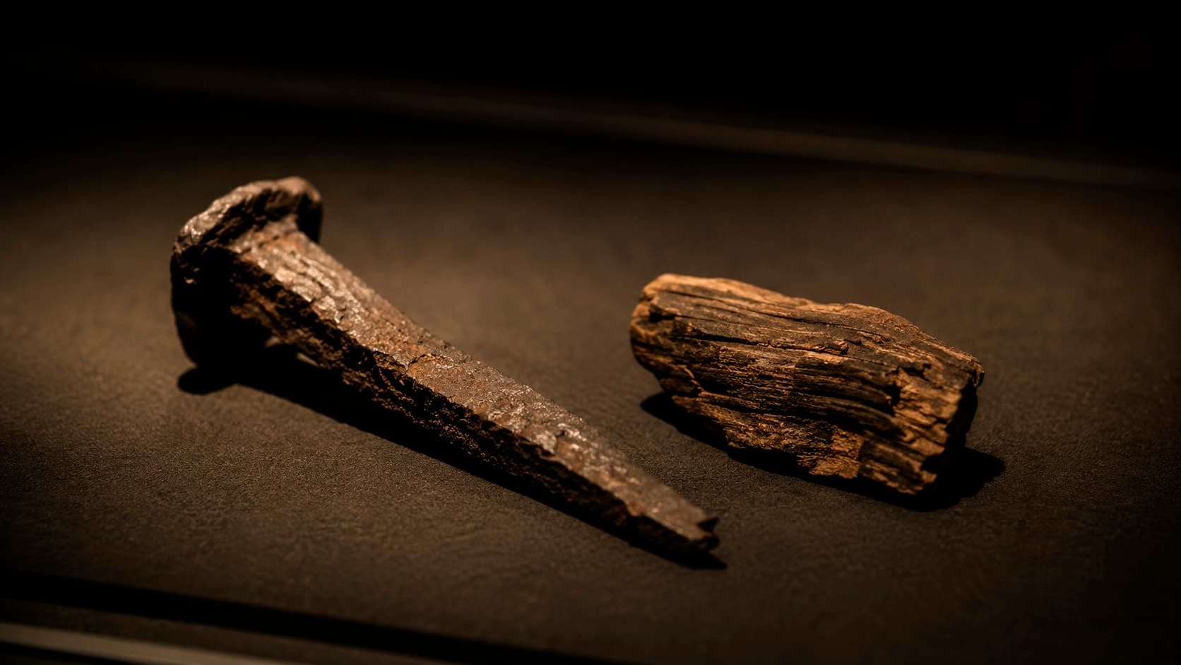 Um prego de ferro antigo e corroído ao lado de um fragmento de madeira desgastada, dispostos sobre superfície escura como artefatos de museu sob luz lateral