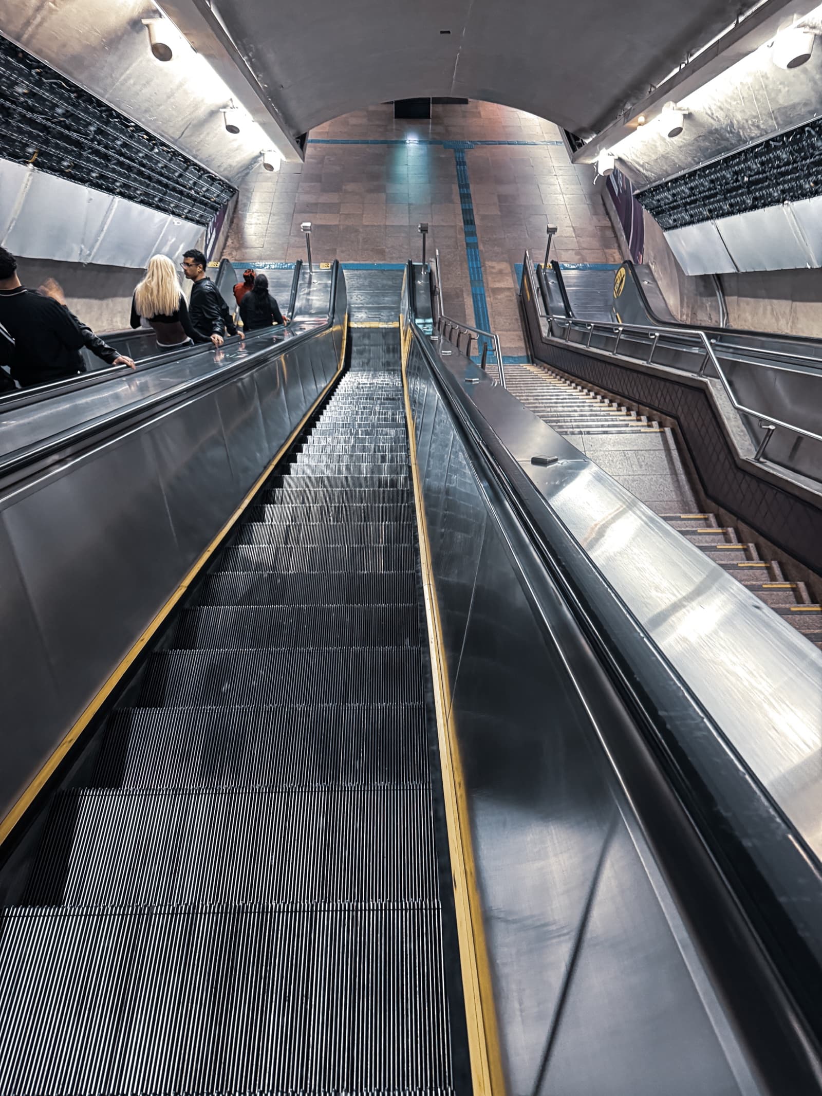 Escada rolante longa e vazia vista de baixo numa estação de metrô, com pessoas de costas na escada ao lado e uma escada fixa à direita
