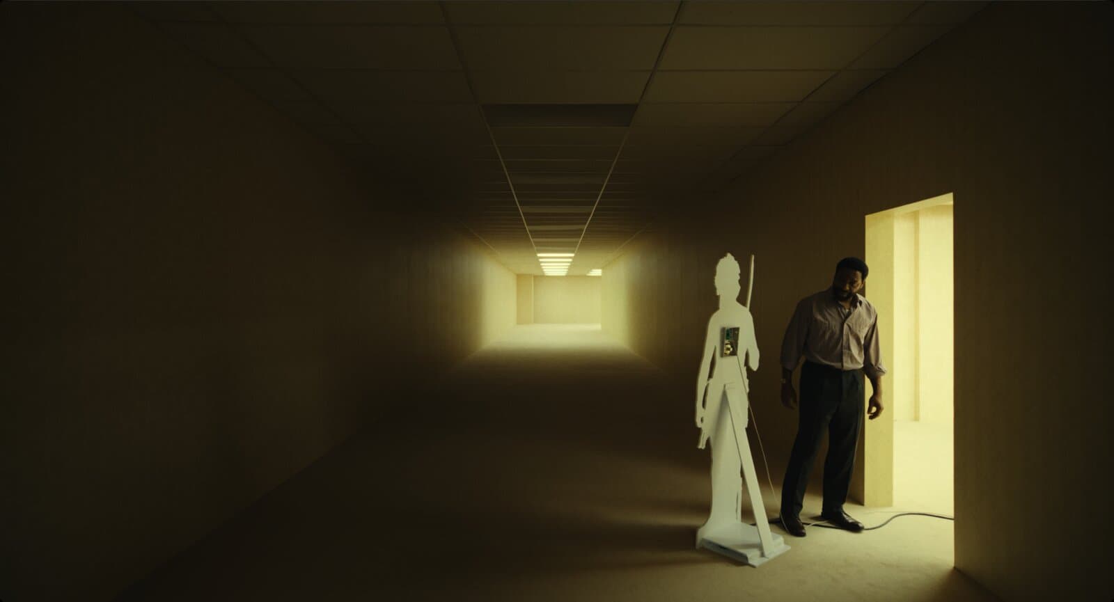 Corredor amarelo vazio e mal iluminado. Perto de uma porta iluminada ao fundo, um homem de camisa está de pé ao lado de um recorte de papelão em tamanho real de um homem das cavernas com uma lança, com um pequeno gravador preso ao peito e um cabo saindo pelo chão.
