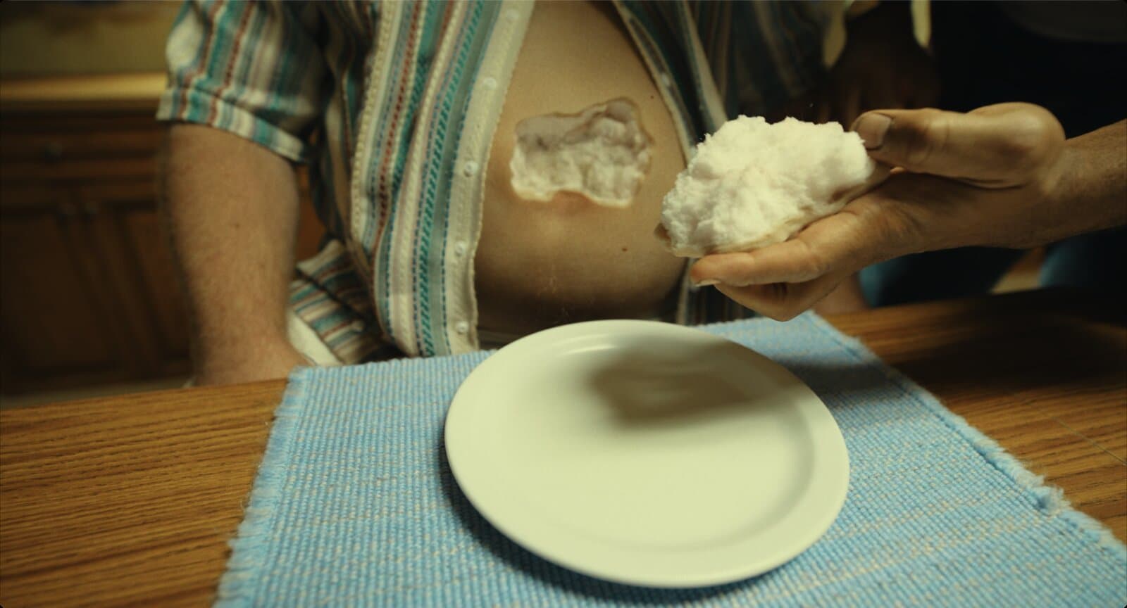 Um homem sentado à mesa, de camisa listrada aberta, tem uma abertura no abdômen preenchida por uma massa branca e esponjosa. Ao lado, uma mão segura um pedaço desse material. Em primeiro plano, um prato branco vazio sobre um jogo americano azul.