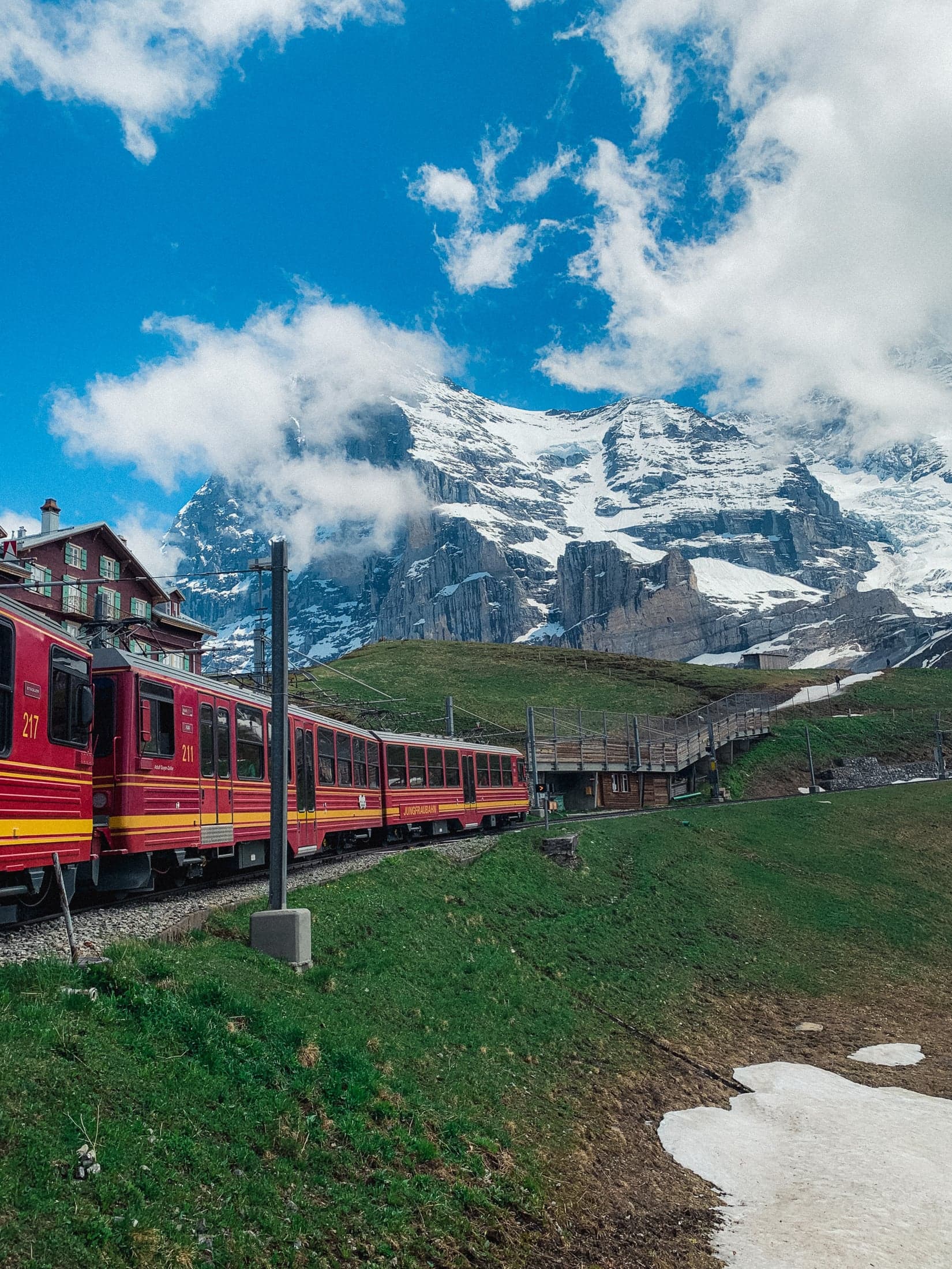 Trem vermelho contornando uma encosta verde, com chalés e uma montanha nevada imponente ao fundo sob nuvens