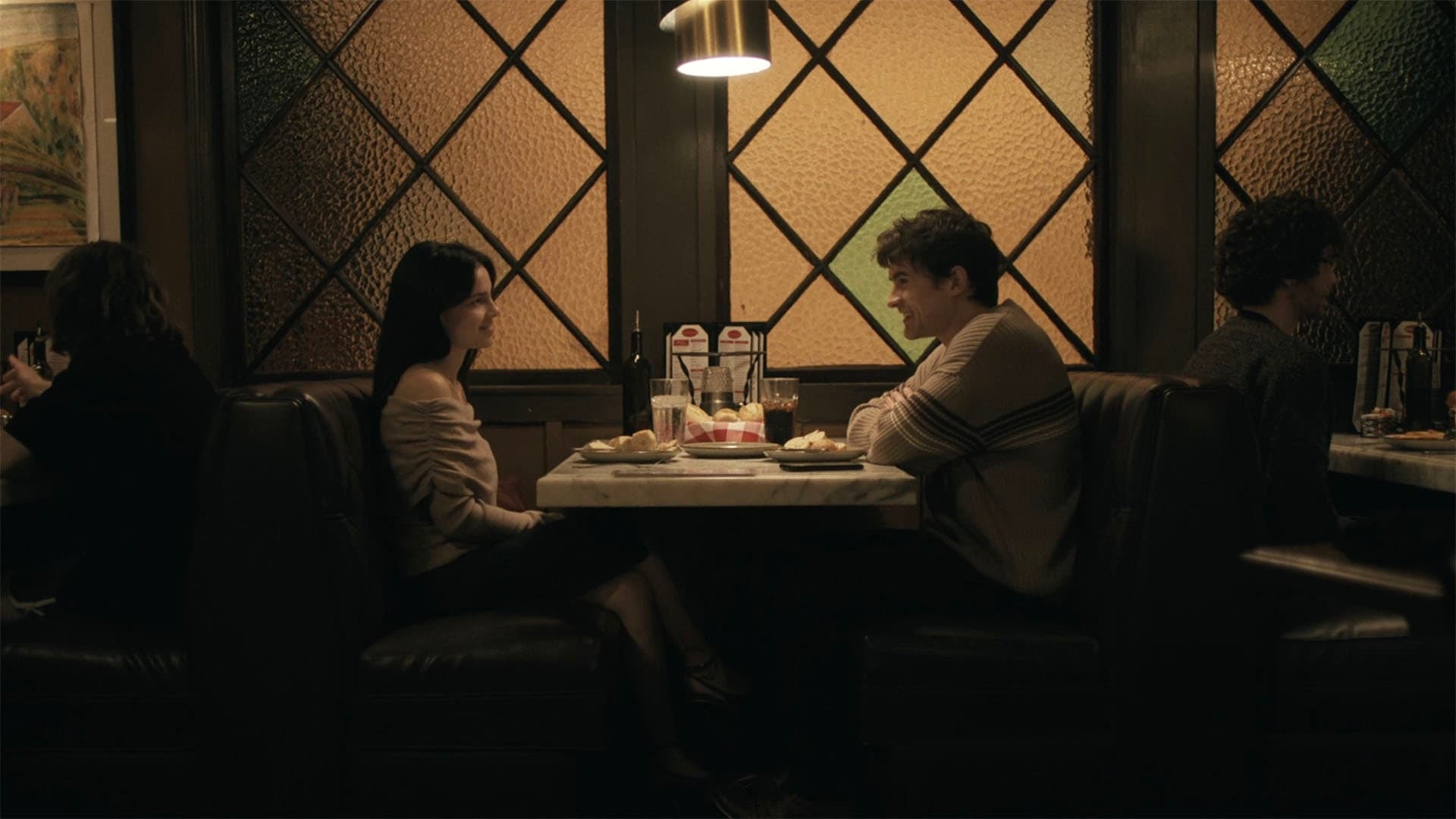 Um homem e uma mulher sentados frente a frente numa mesa de lanchonete, à luz baixa, vistos de lado