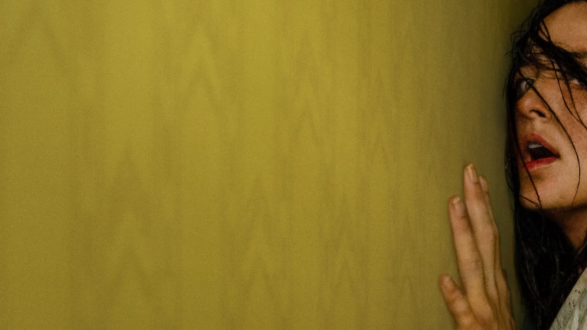 Uma grande parede amarela vazia ocupa quase todo o quadro; no canto direito, cortado pela borda, o rosto de uma mulher de boca aberta, com a mão espalmada contra a superfície.
